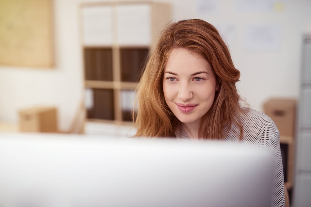 Young woman working on a desktop computer smiling as she leans forwards reading text on the screen, view over the monitor