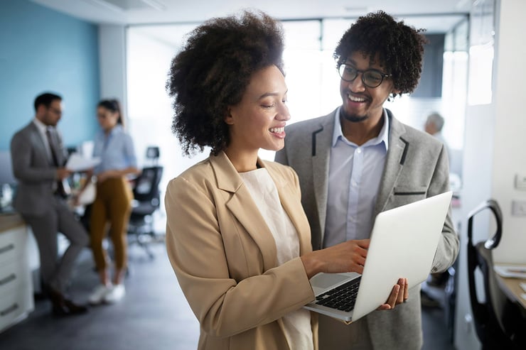 A female employee is seen standing, holding her work laptop, and showing her coworker something new she just learned about avoiding phishing scams.