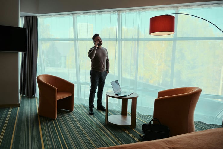 A male sales employee, standing by the window in his hotel room during vacation, takes a client call on his mobile device.