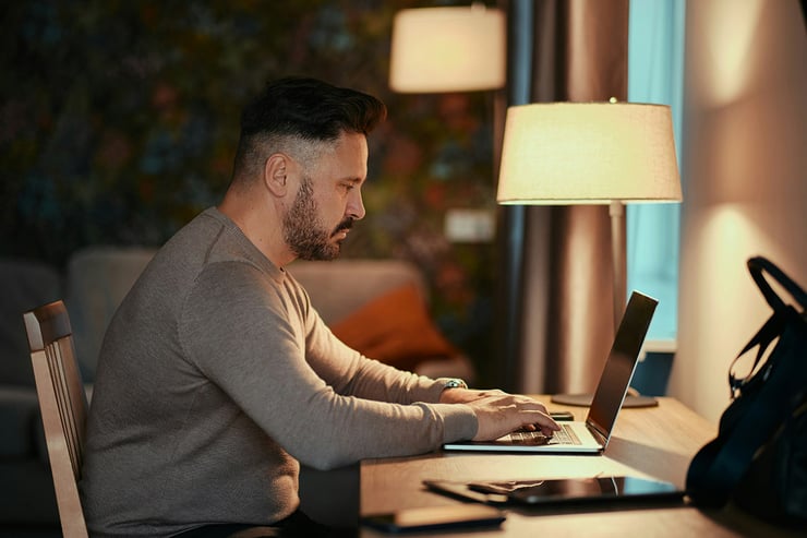 In his hotel room during vacation, a male sales employee sits at his desk using Microsoft Outlook on his laptop to address an urgent client need.