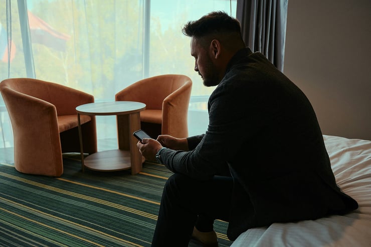 A male sales employee is seen sitting on his hotel bed during vacation, fully plugged into work through his device.