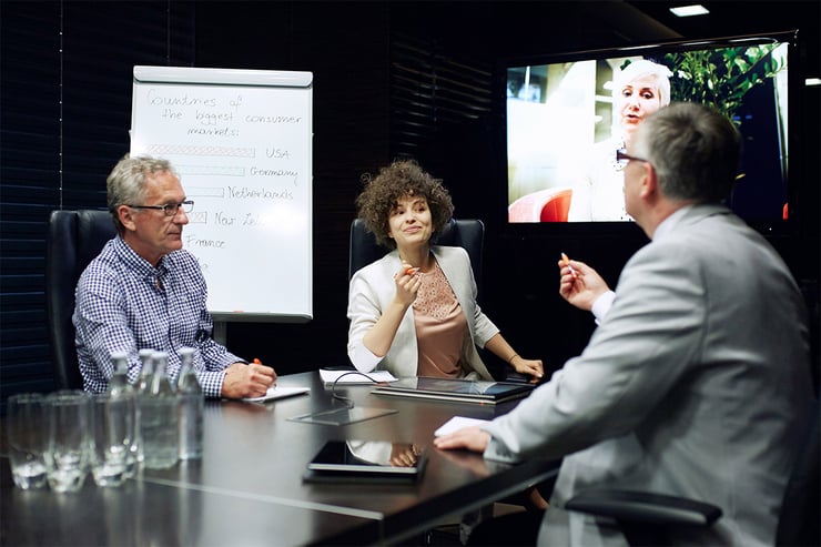 A team at an accounting firm is meeting with remote and on-site members to discuss their major upcoming report where they plan to present it to their client using a variety of technologies such as tablets, large monitors, and laptops, which facilitate note-sharing, video calls, emails, and document handling.