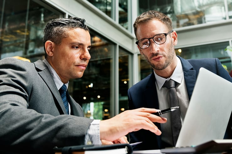 Two male employees at an insurance company meet in their office lobby, intently reviewing a PowerPoint presentation on a laptop, highlighting a common challenge in the finance industry: enabling secure, seamless collaboration and access to critical files in a fast-paced, highly regulated environment.