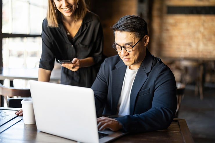 A male customer success manager from a financial services organization types on his work laptop, collaborating with his team on a shared document to strengthen client relationships, while another employee smiles with a mobile work device, highlighting how IT support for financial services enables secure collaboration, seamless tool access, and reliable connectivity to drive productivity and business growth.