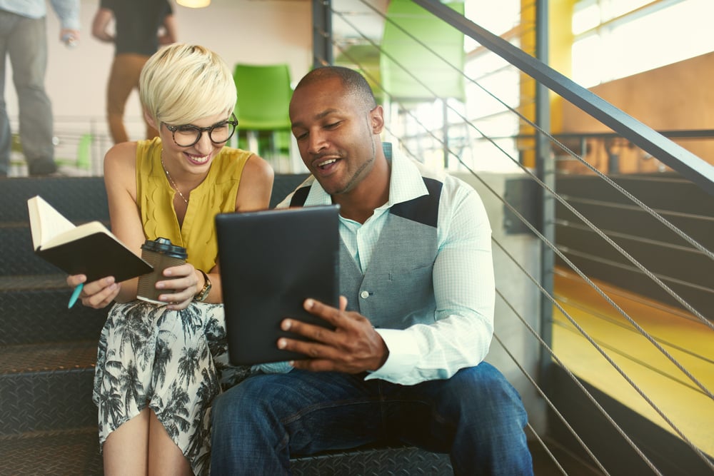 Two creative millenial small business owners working on social media strategy using a digital tablet while sitting in staircase-1