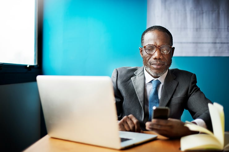 A male employee sits at his office desk with his laptop open, holding his work mobile device as he scrolls through missed messages in Microsoft Teams, effortlessly staying connected and catching up on important conversations with his team.