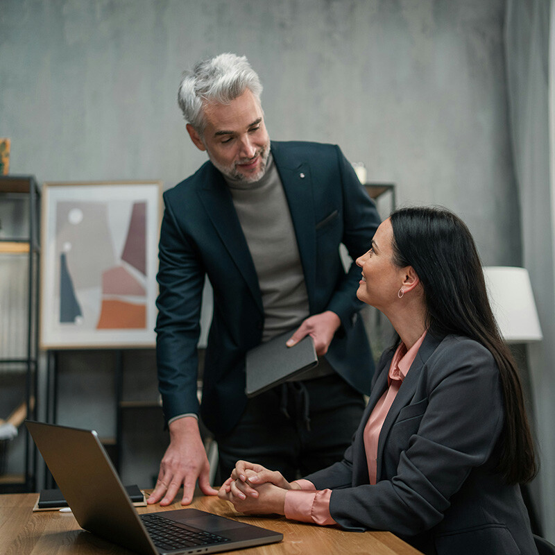 A manager and an employee in an office look at the employee's laptop screen, which shows 